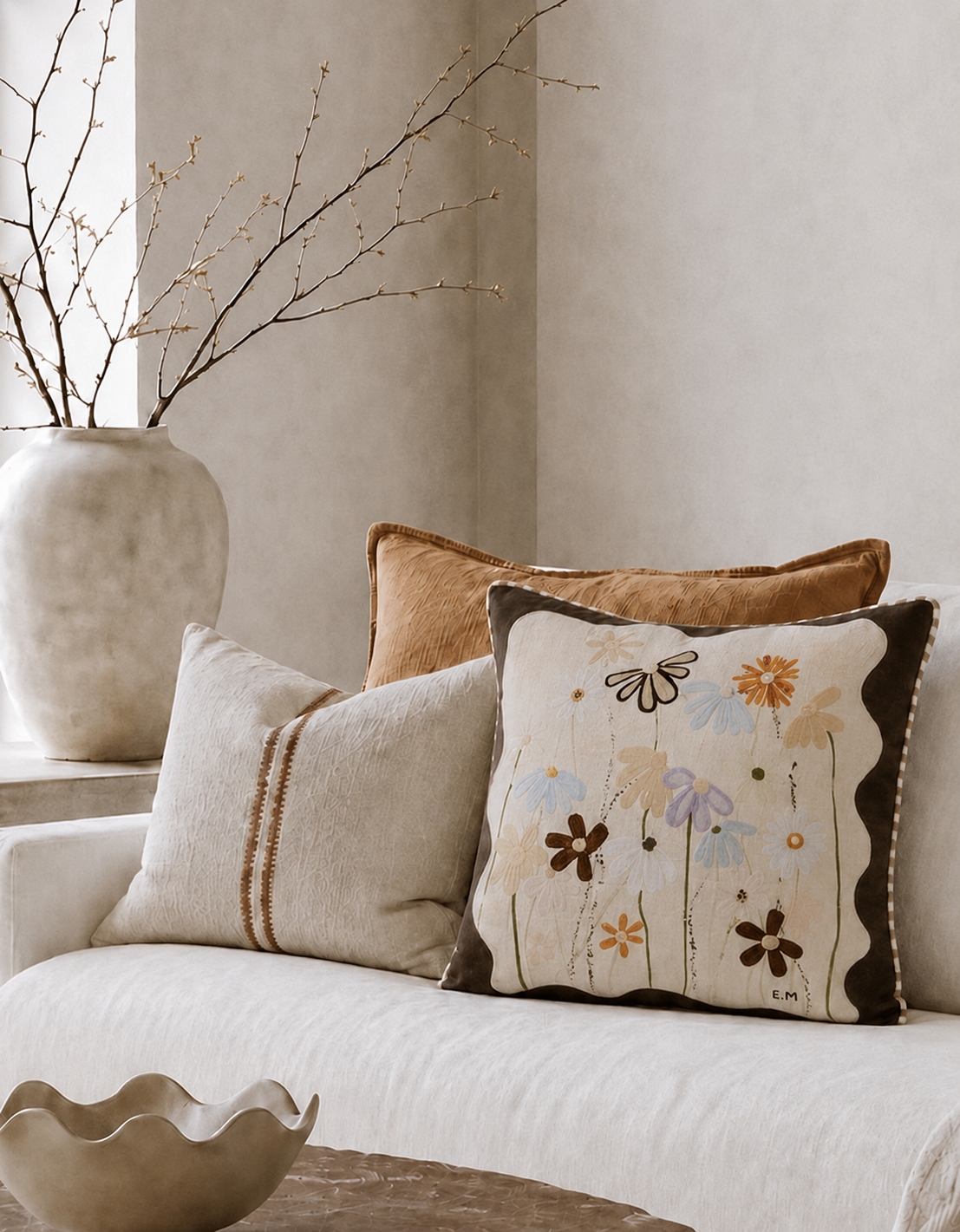 A styled interior scene with a white sofa holding three decorative throw pillows. The front pillow is cream with embroidered floral and butterfly motifs and dark brown border details; the middle pillow is light beige with brown stripe accents; the back pillow is a warm tan/brown.