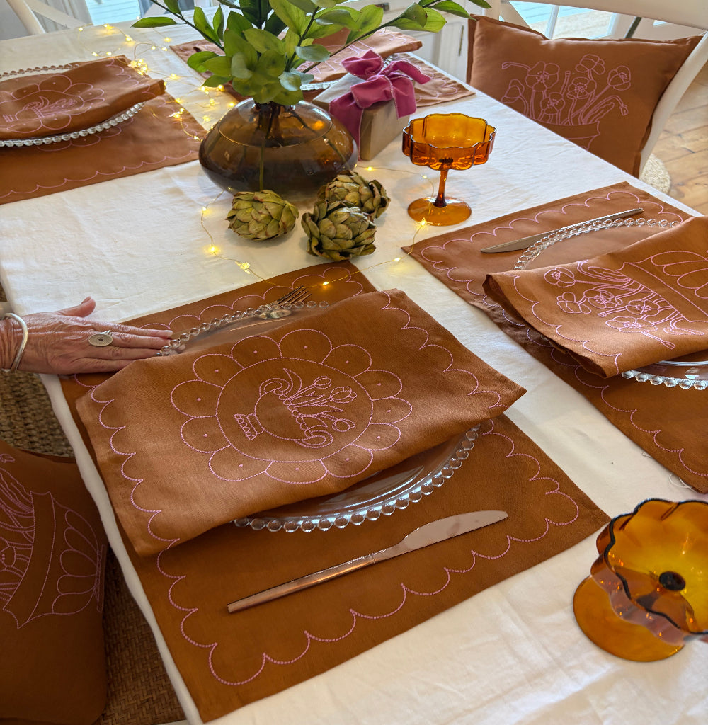 Decorative table setting with tan and pink placemats and cutlery on a white tablecloth.