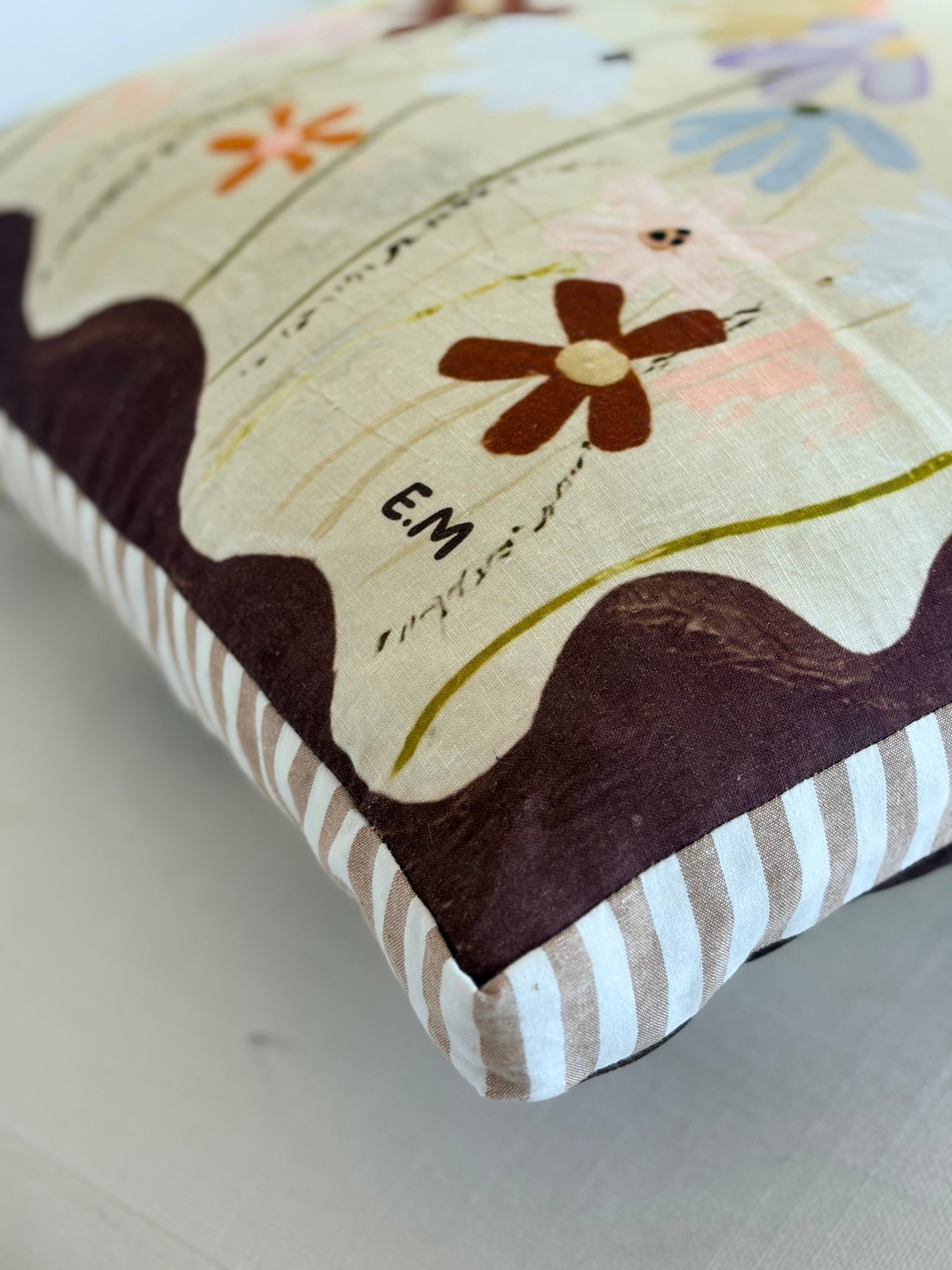 A close-up of a decorative throw pillow with a cream background, dark brown abstract shapes, a brown flower motif, and a striped border in beige and white.
