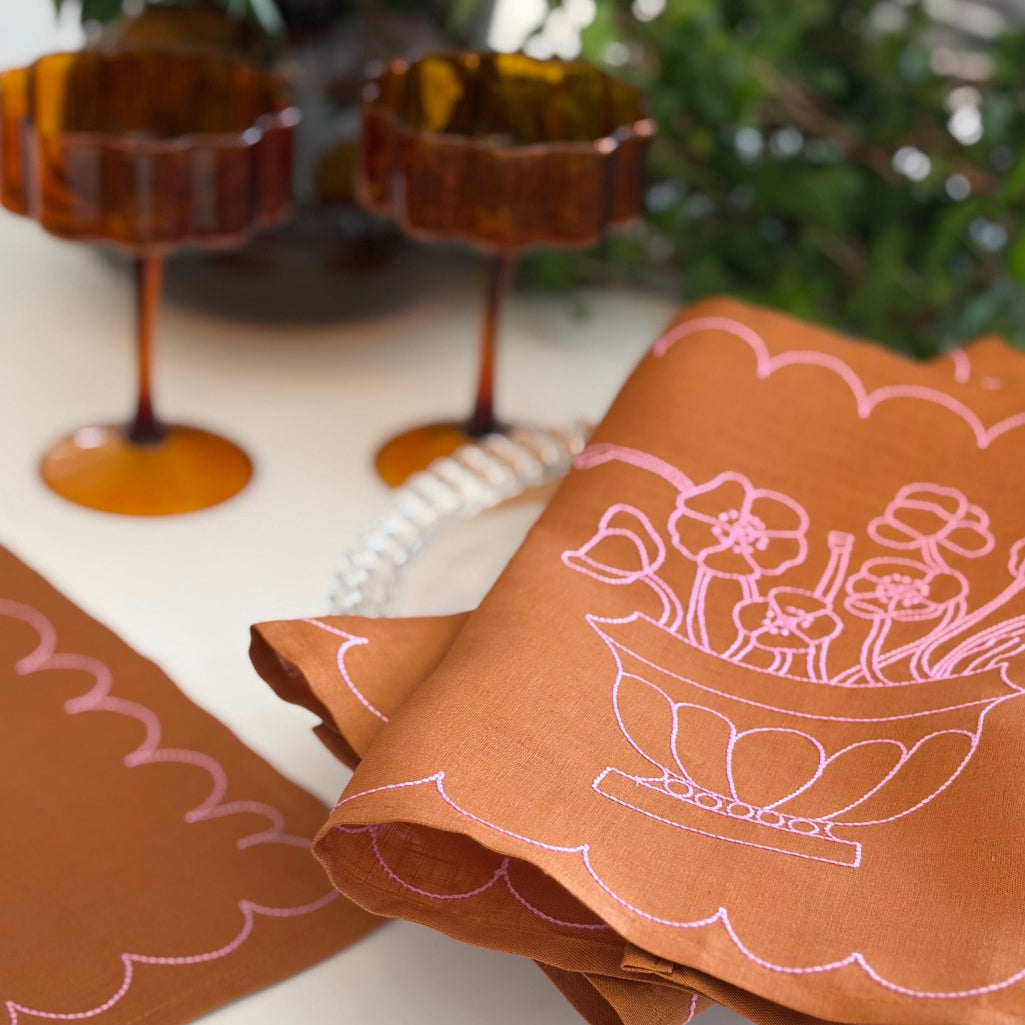 Tan linen fabric placemat with pink embroidery on a table with decorative items in the background