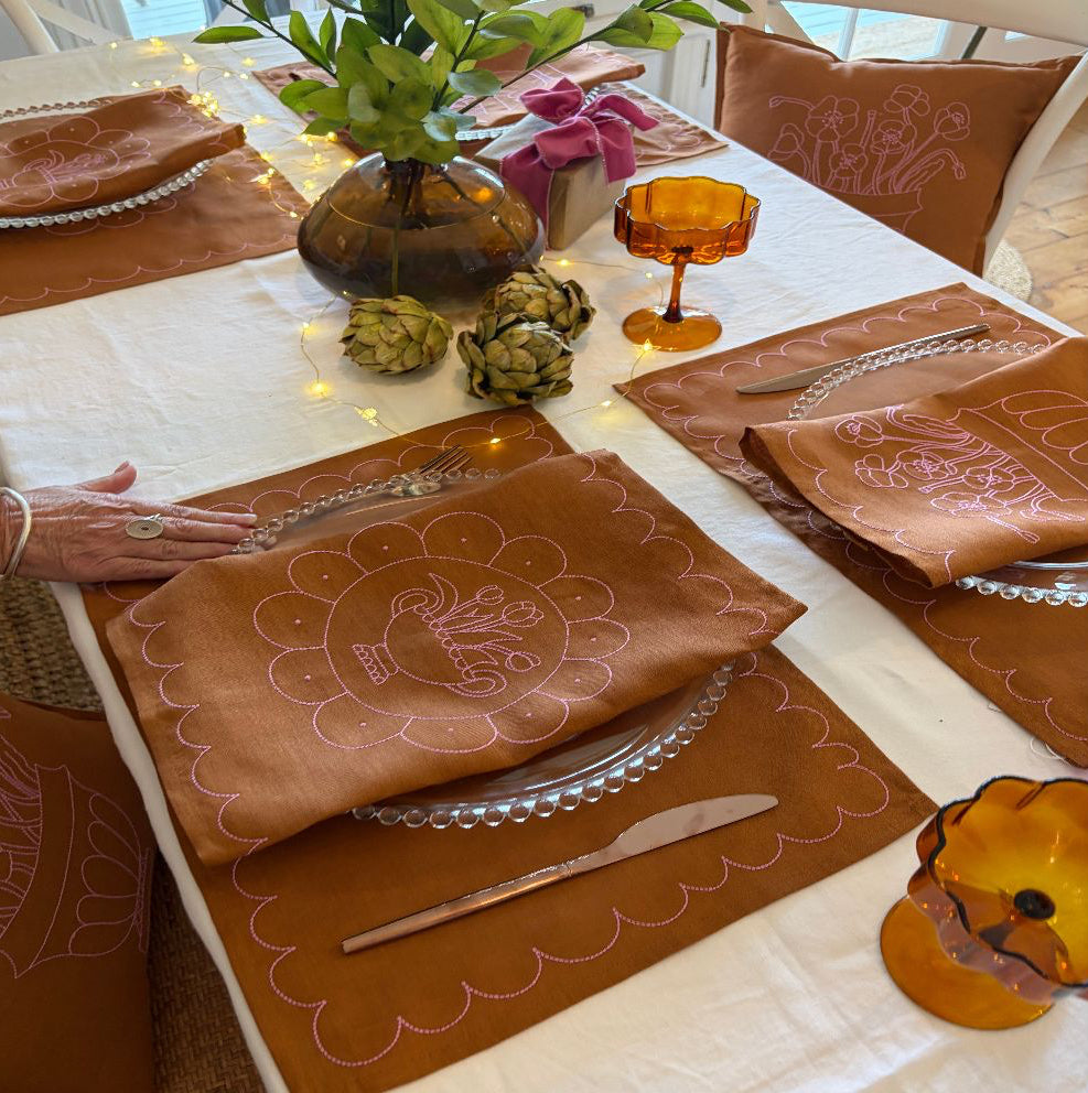 Decorative table setting with tan and pink placemats and cutlery on a white tablecloth.
