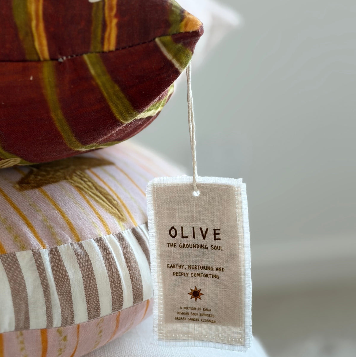 A close-up of a striped decorative pillow with a hanging fabric tag that reads 'OLIVE THE GROUNDING SOUL' and 'EARTH, SUSTAINABLE AND DELICIOUSLY COMFORTING.'.