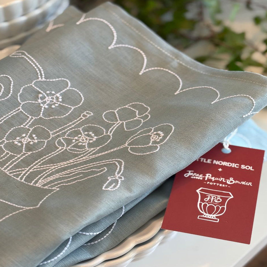 Blue and white linen placemat with floral poppies  embroidery and a brand tag  Janae Paquin Bowden and Little Nordic Sol on a white surface