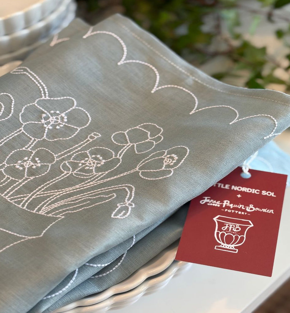 Blue and white linen placemat with floral poppies  embroidery and a brand tag  Janae Paquin Bowden and Little Nordic Sol on a white surface