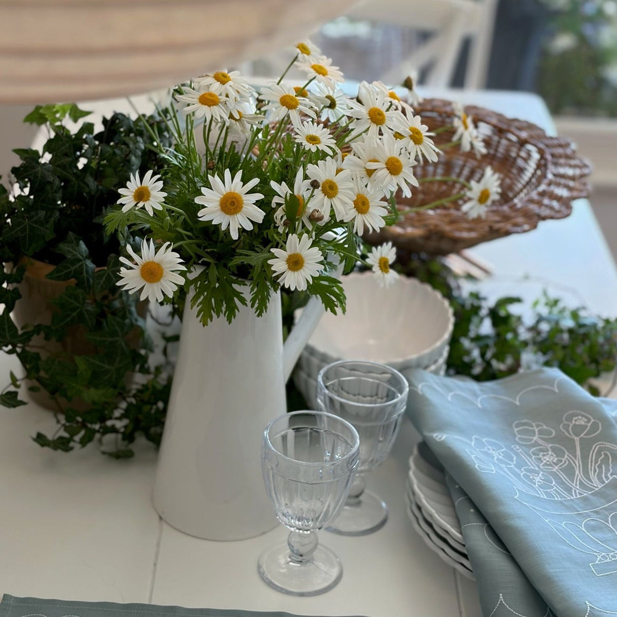 White pitcher with flowers on a table setting with glasses and plates.
