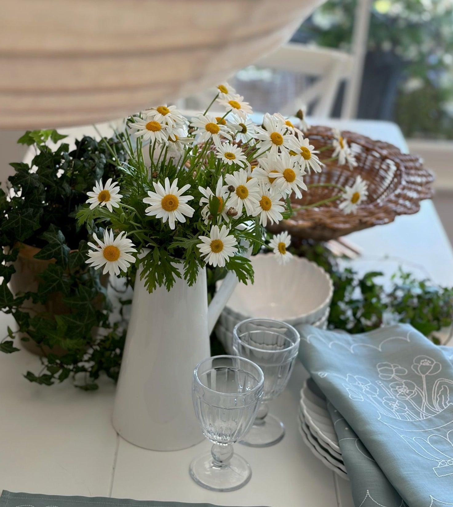 White pitcher with flowers on a table setting with glasses and plates.