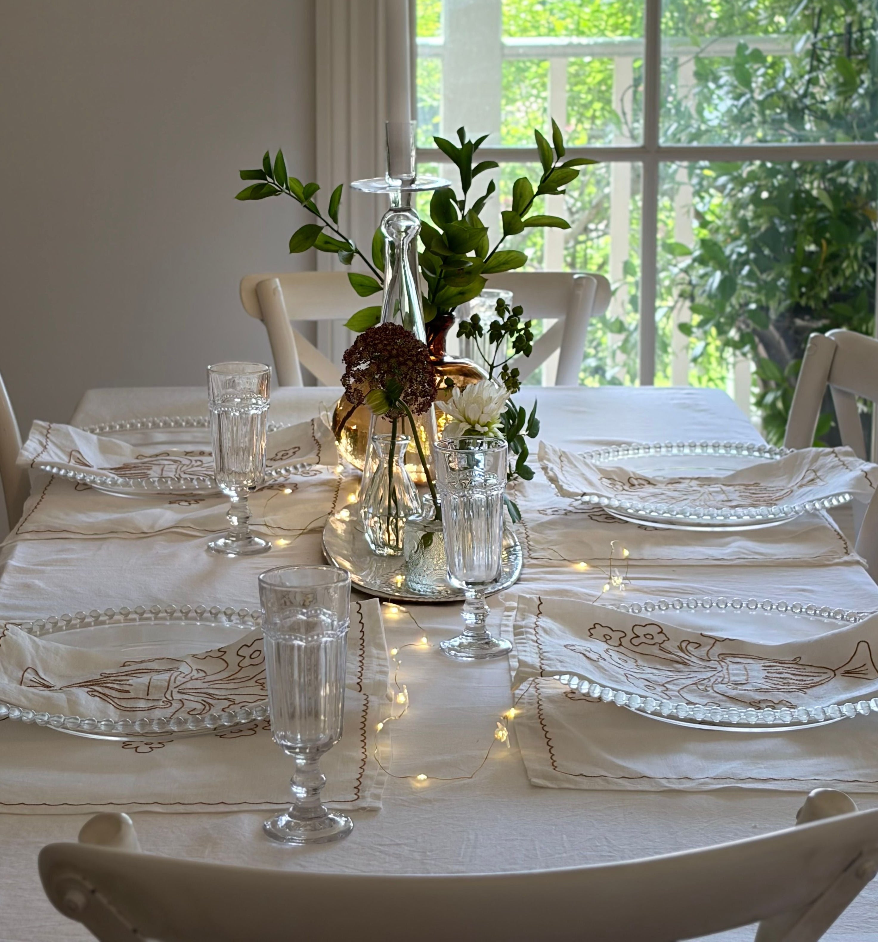 Elegant table setting with white tablecloth, glassware, and decorative centerpieces in a bright room.