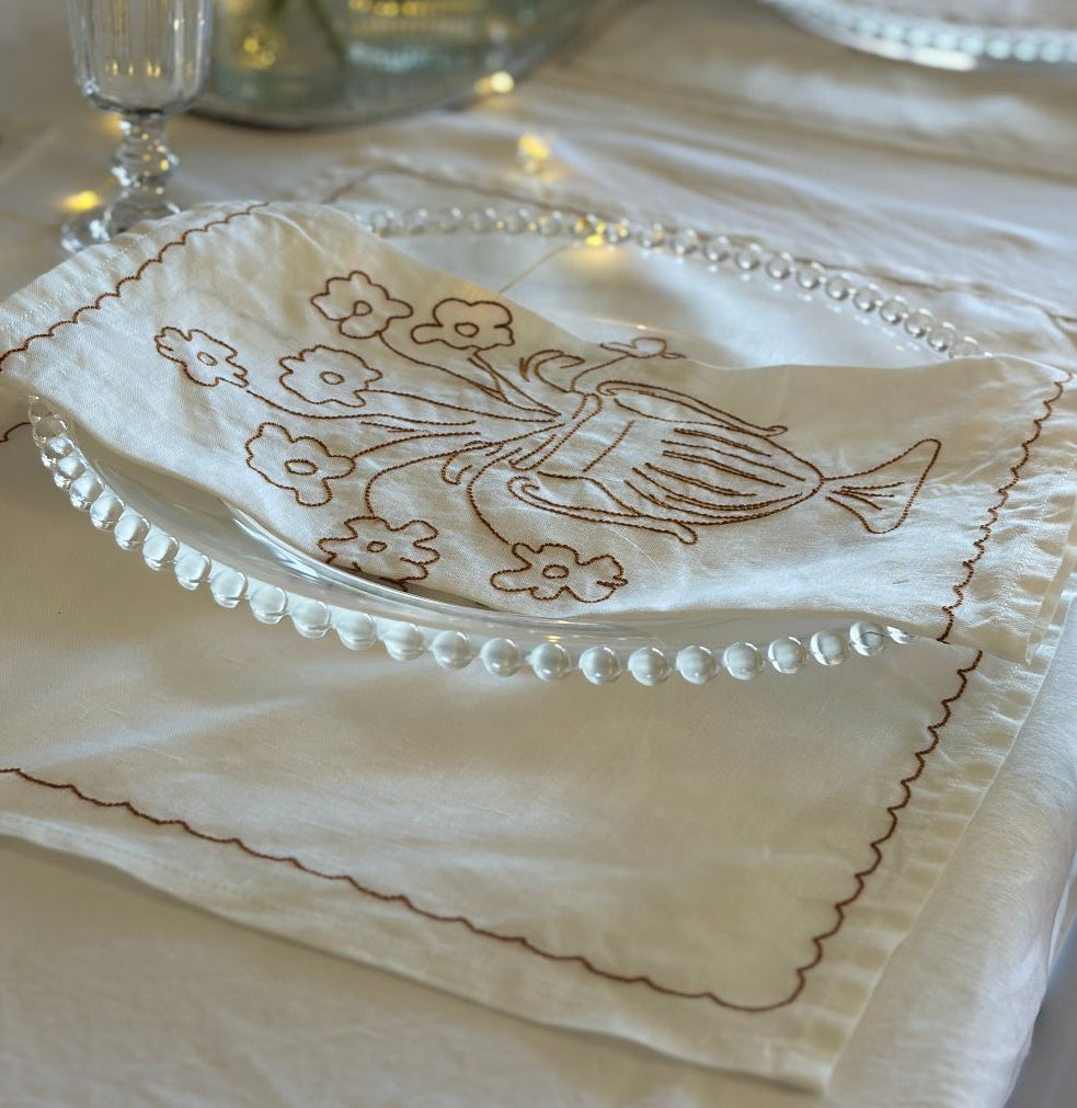 Embroidered napkin with floral design on a glass plate over a white tablecloth.