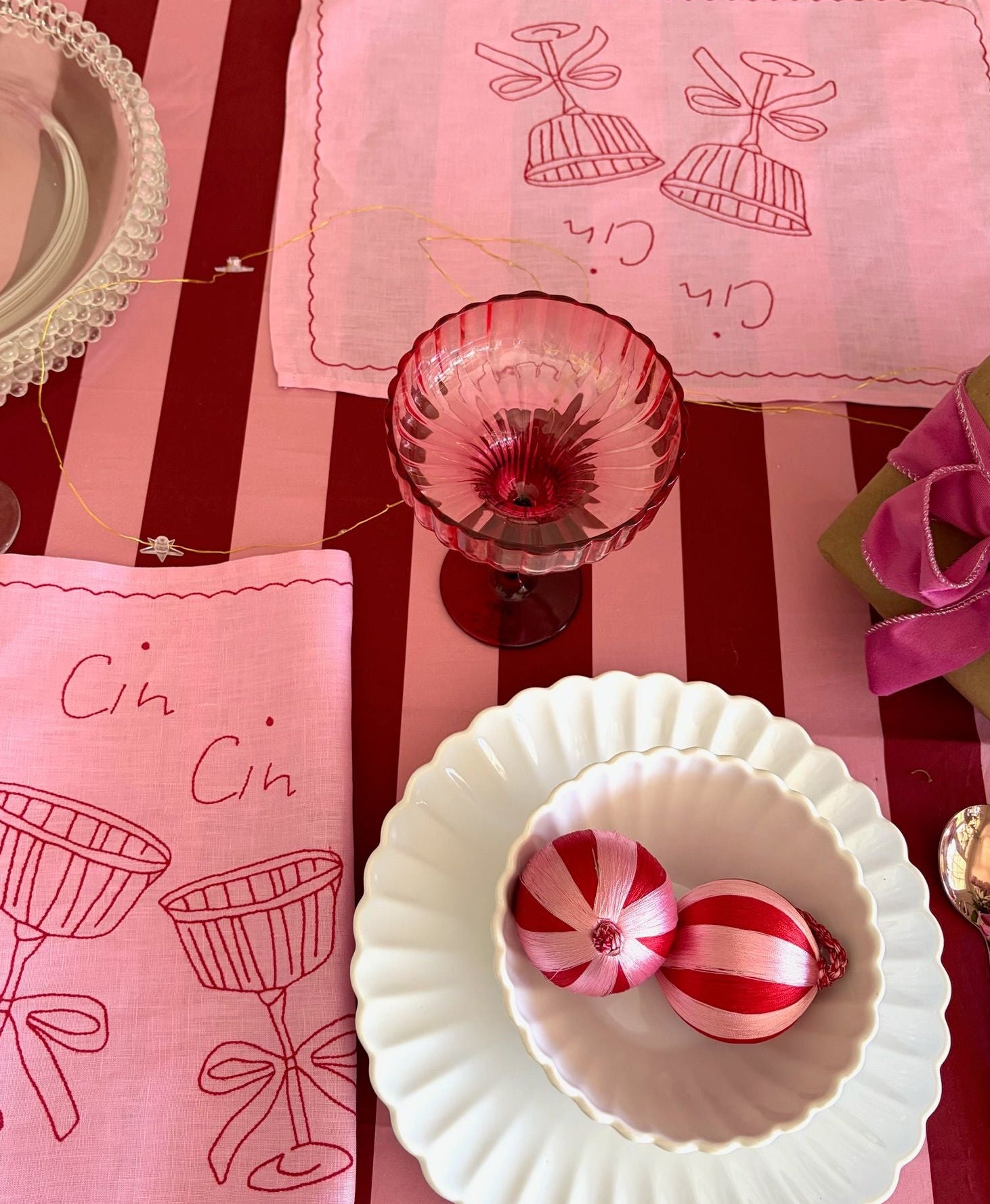 Table setting with pink and red accents, including a glass, plates, and decorative items on a striped tablecloth.