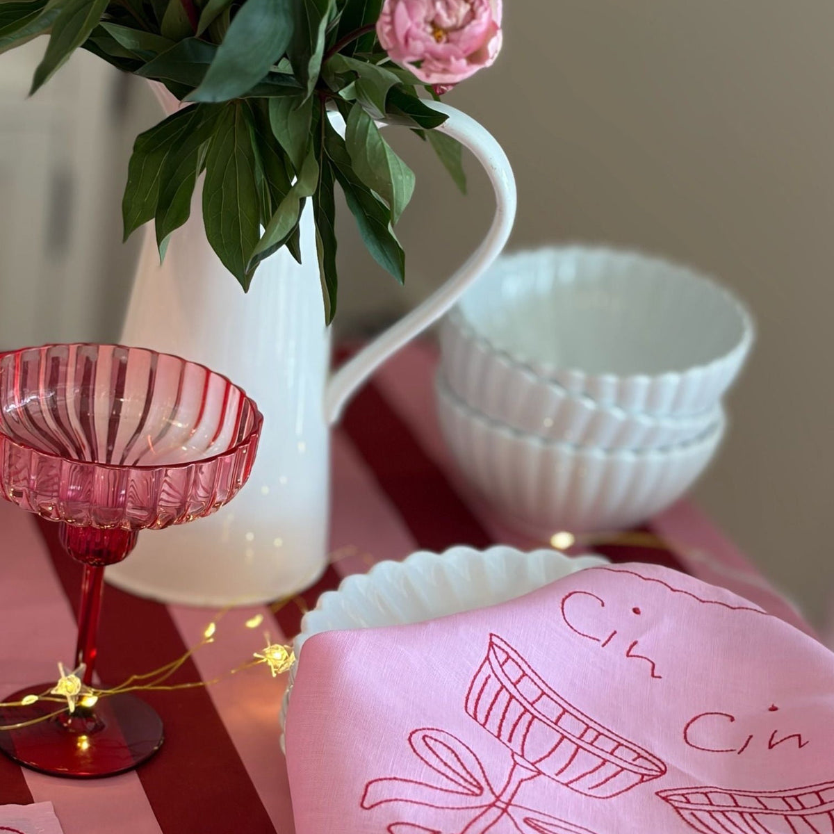 Pink tablecloth with 'Cin Cin' design, red glass, white vase with flowers, and white bowls on a table.