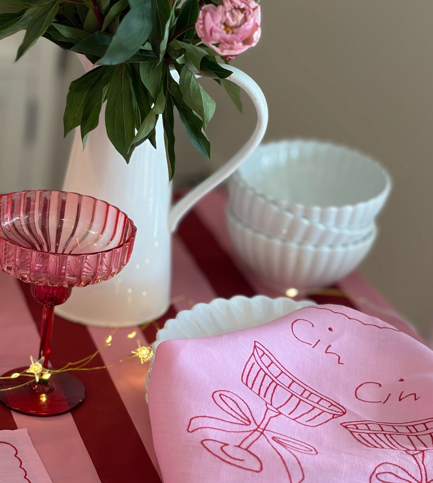 Pink tablecloth with 'Cin Cin' design, red glass, white vase with flowers, and white bowls on a table.
