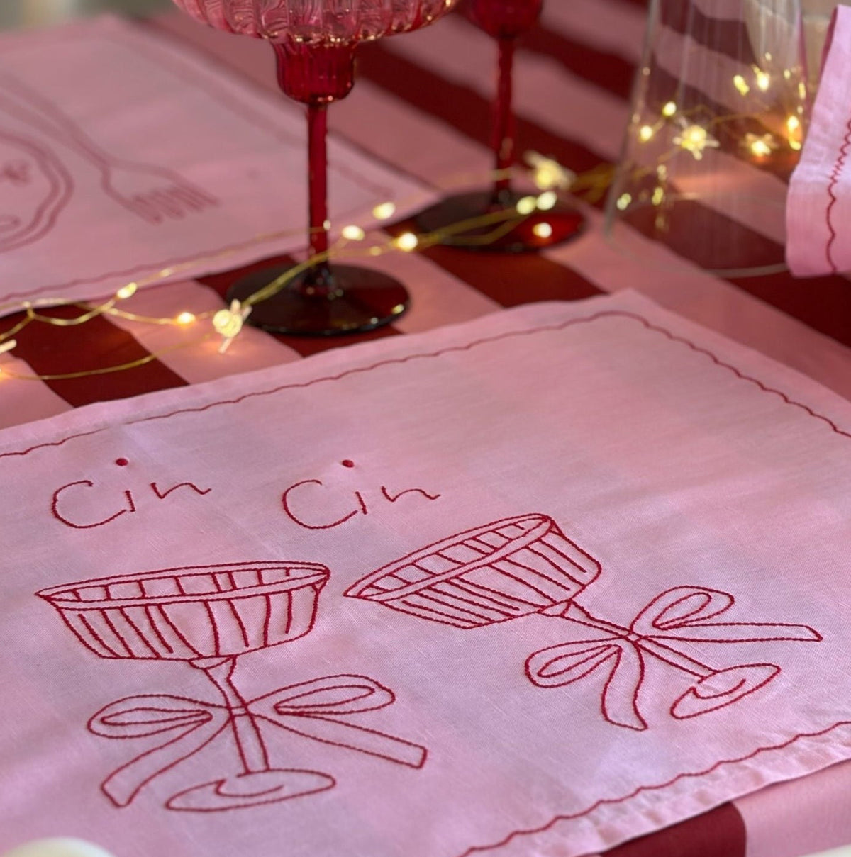 Pink glasses and embroidered towel with 'Cin Cin' design on a table.