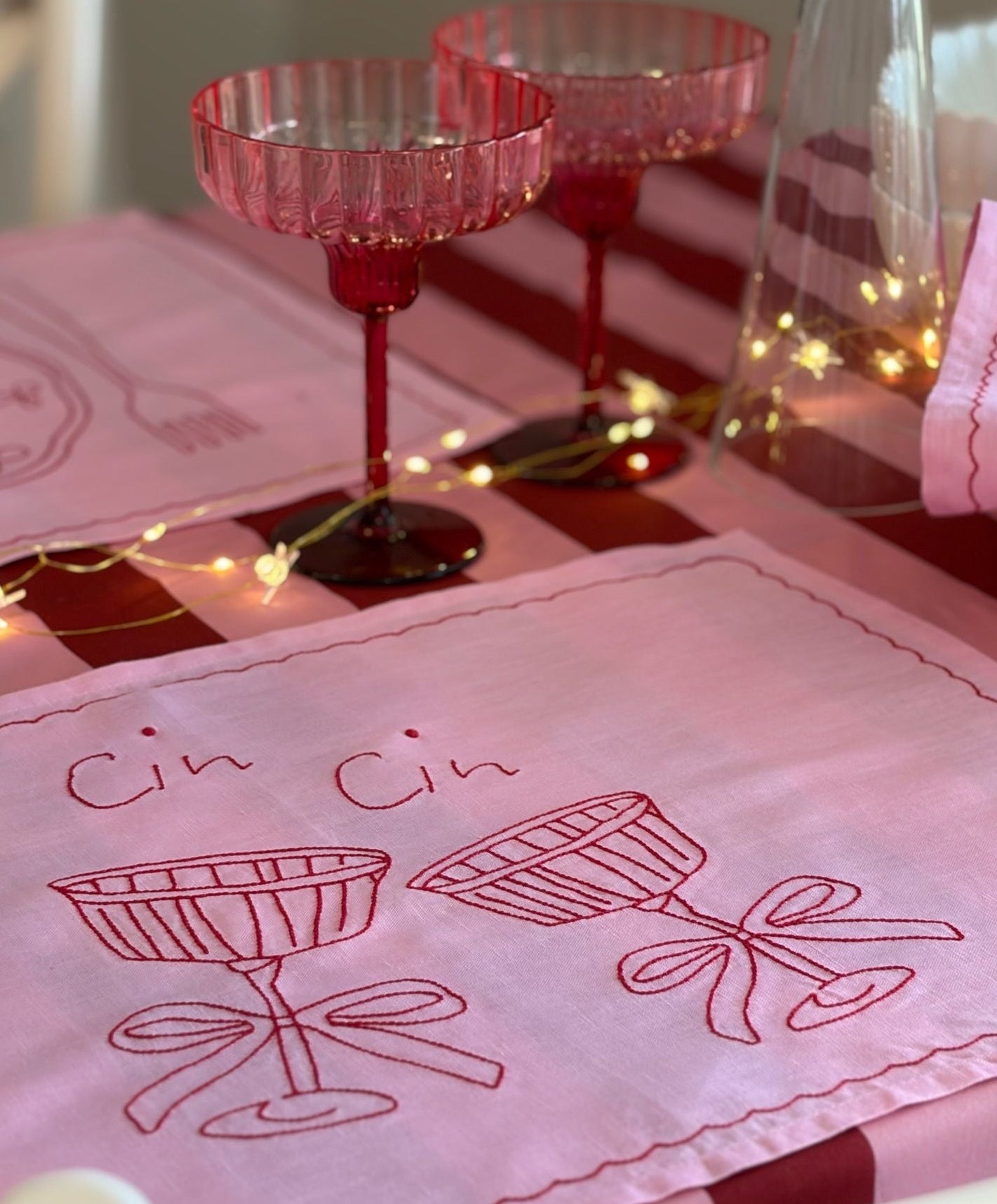 Pink glasses and embroidered towel with 'Cin Cin' design on a table.