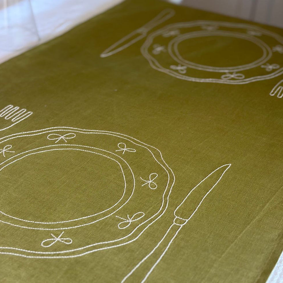 Close-up of Little Nordic Sol Duo olive linen placemat featuring white embroidered plates and cutlery.