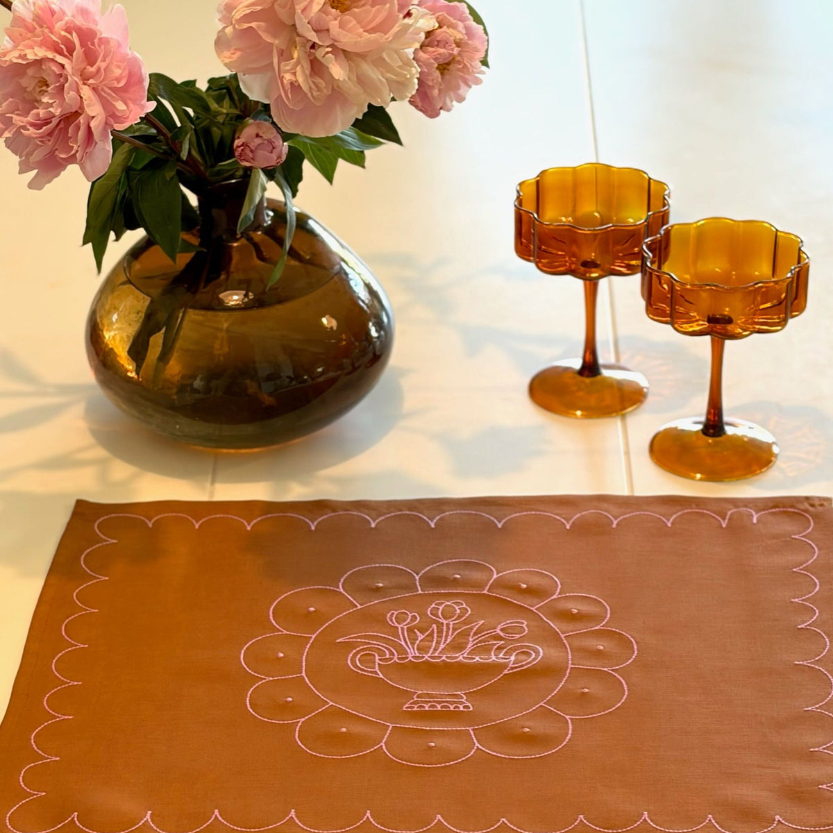Decorative table setting with a vase of pink flowers, two amber glasses, and a brown placemat with floral design.