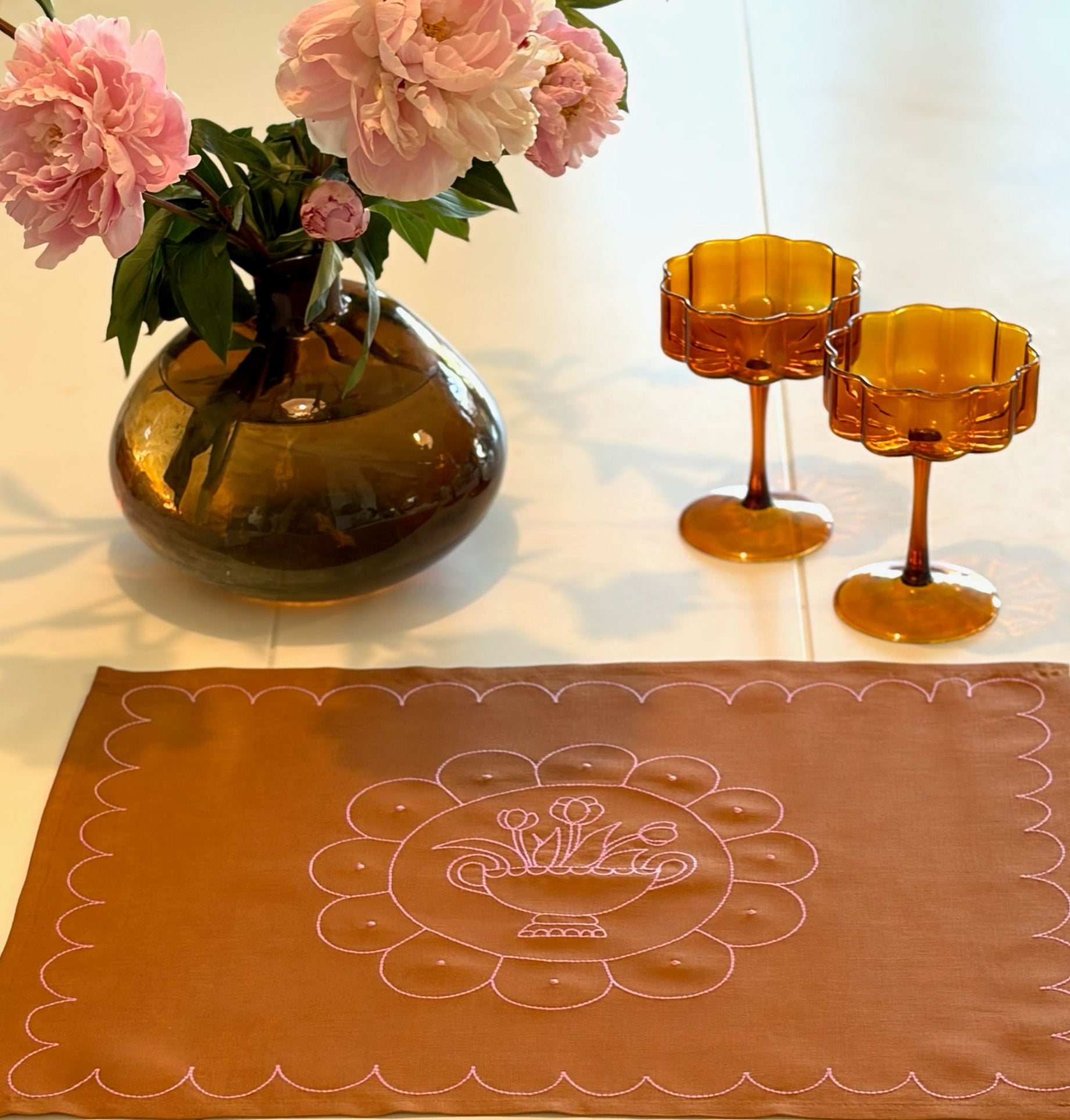 Decorative table setting with a vase of pink flowers, two amber glasses, and a brown placemat with floral design.