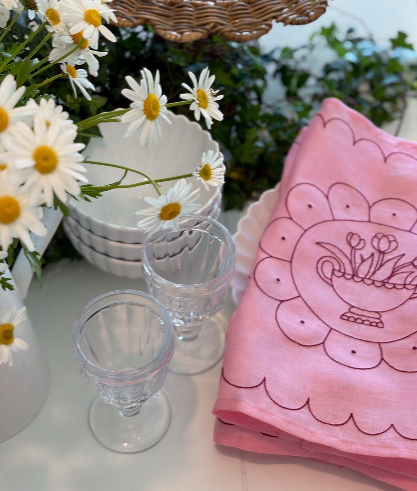 Pink embroidered napkin with floral design on a table with white bowls, glasses, and daisies.