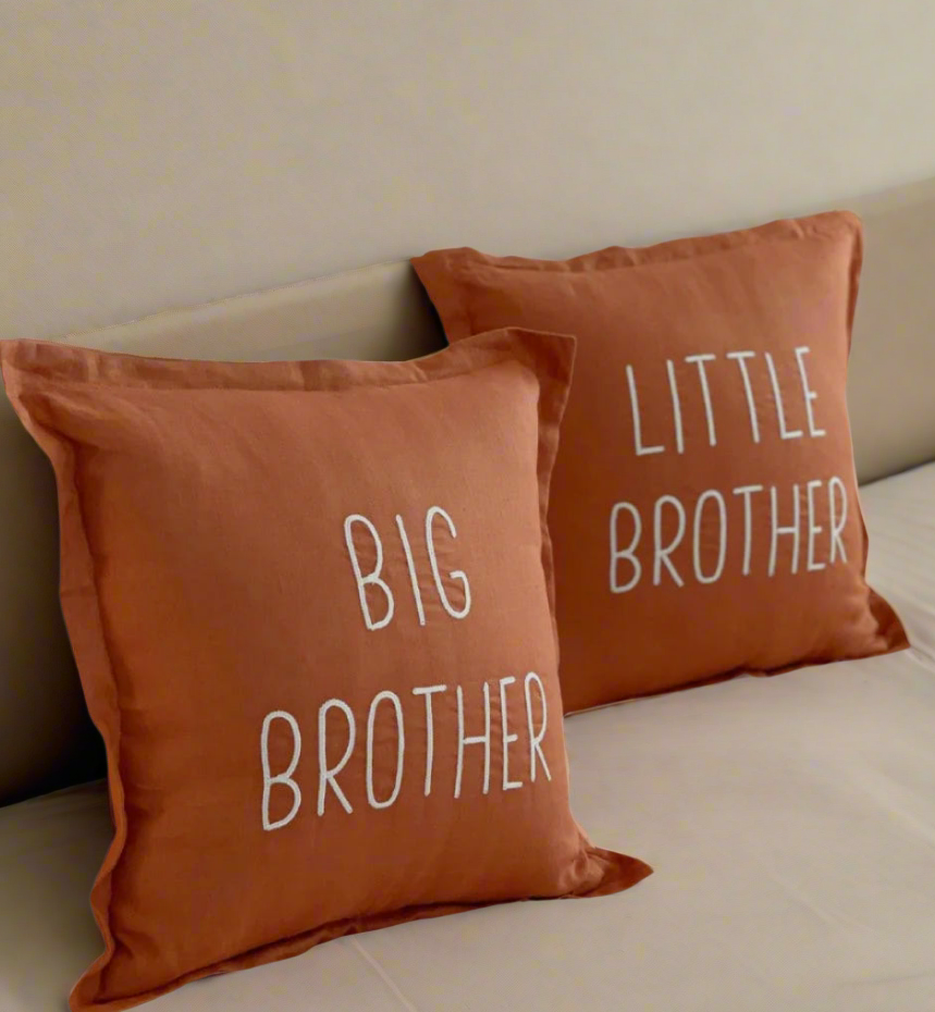 Two rust coloured pillows with 'Big Brother' and 'Little Brother' text on a bed.