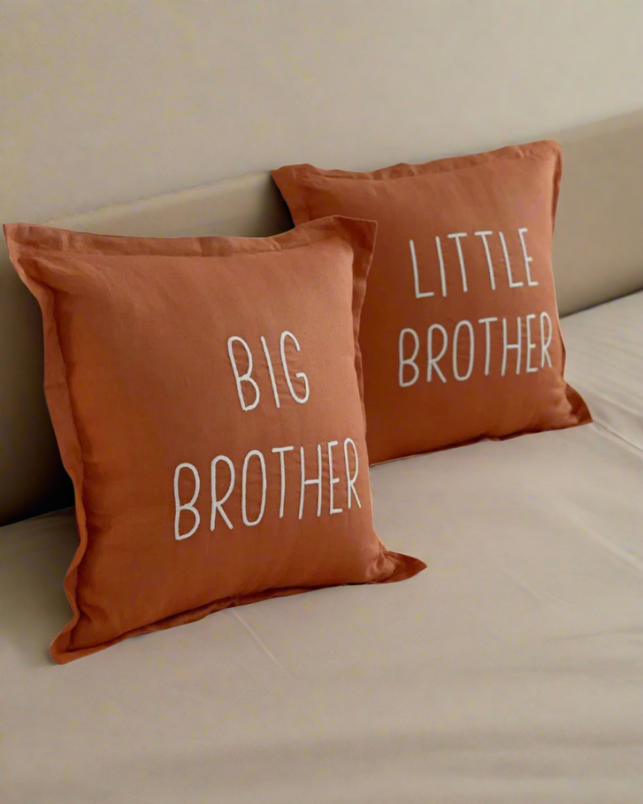 Two rust coloured pillows with 'Big Brother' and 'Little Brother' text on a bed.