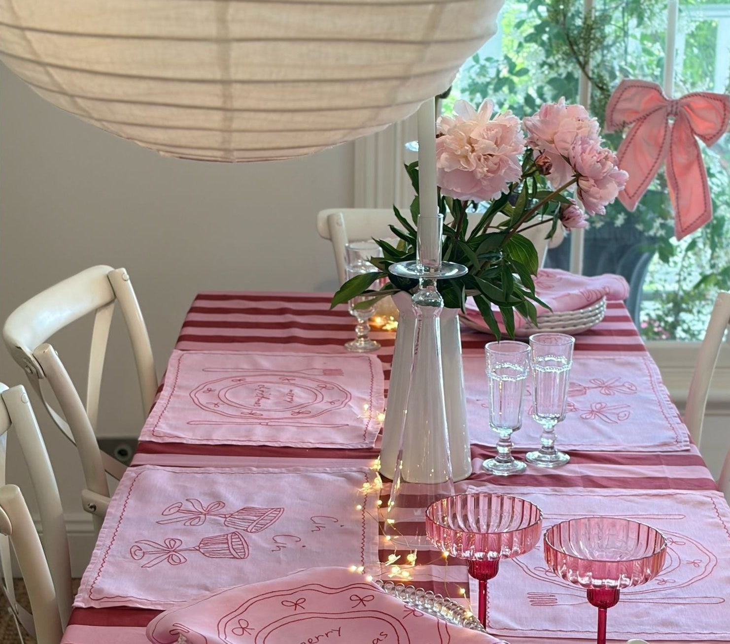 Dining table set with pink tablecloth, glasses, and flowers in a bright room.